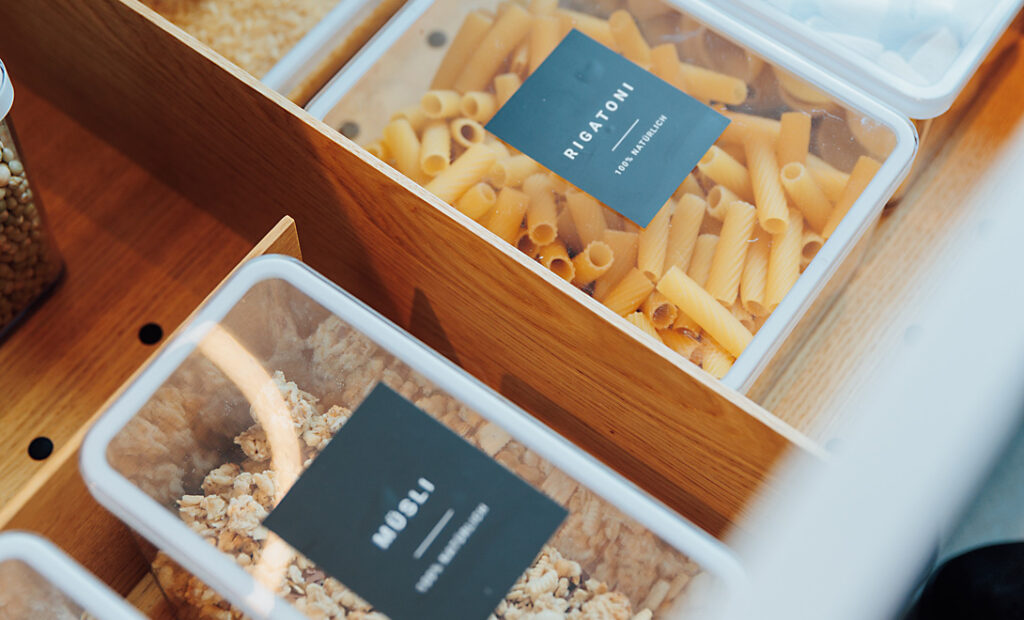 Holzschublade mit durchsichtigen Boxen, ordentlich gefüllt und beschriftet: Rigatoni, Müsli, Reis und weitere Vorräte.