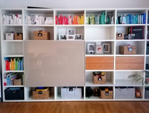 Weißes Regal mit Büchern nach Farben sortiert, Körben, Bildern und Spielzeugkisten.