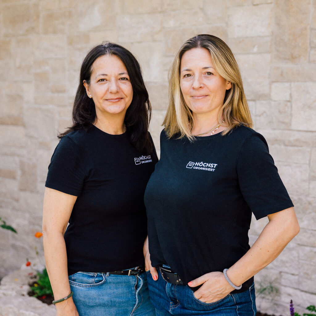 Zwei Frauen im schwarzen „Höchst Organisiert“-Shirt stehen nebeneinander lächelnd vor einer hellen Steinwand im Freien.
