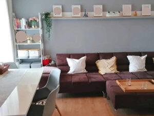 Wohnzimmer mit braunem Sofa, Tisch voller Gegenstände und Regal im Hintergrund – sichtbare Unordnung.
