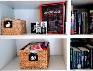 Regal mit Büchern und Depeche-Mode-Fanartikeln, dekorative Körbe mit Symbolschildern.