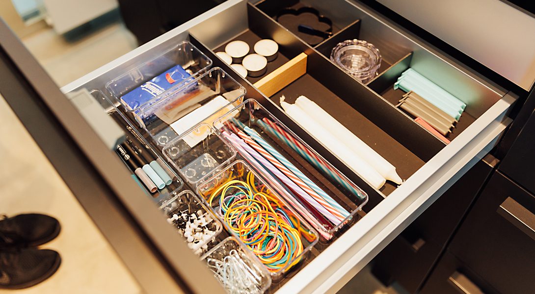 Geöffnete Schublade mit transparenten Boxen, sortiert gefüllt mit Gummibändern, Kerzen, Clips, Stiften und kleinen Utensilien.