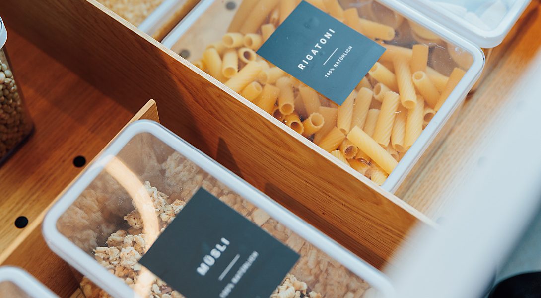 Holzschublade mit durchsichtigen Boxen, ordentlich gefüllt und beschriftet: Rigatoni, Müsli, Reis und weitere Vorräte.