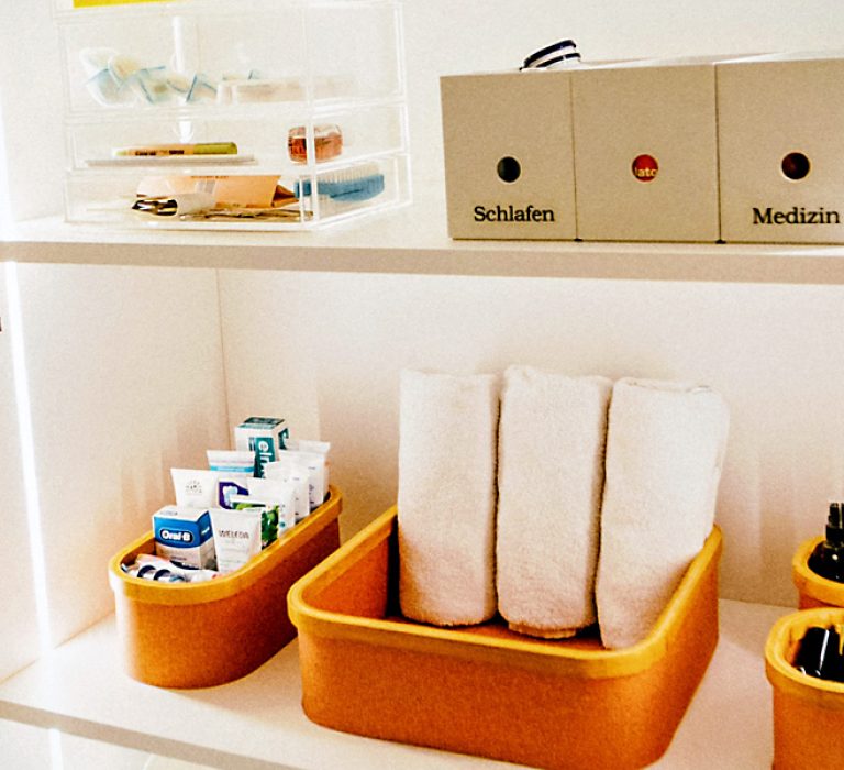 Offener Schrank mit gelben Boxen, gerollten Handtüchern, Pflegeprodukten und beschrifteten Ablagen „Schlafen“ und „Medizin“.
