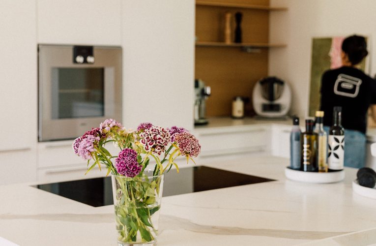 Weiße Küche mit moderner Insel, Glasvase mit lila Blumen im Vordergrund und Regal mit Küchengeräten im Hintergrund.
