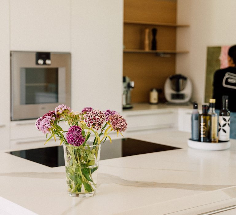Weiße Küche mit moderner Insel, Glasvase mit lila Blumen im Vordergrund und Regal mit Küchengeräten im Hintergrund.