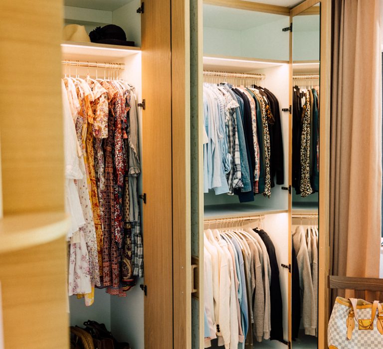 bild_rezensionen_michael Offener Kleiderschrank mit ordentlich sortierten Kleidern, Hemden und Jacken, daneben Fenster mit Pflanze.
