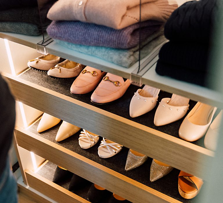 bild_rezensionen_schuhschrank Ordentlich sortierte Regalböden mit flachen Damenschuhen und gefalteten Pullovern in sanften Farben, dezent beleuchtet.