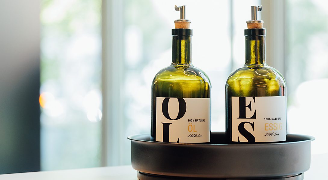 Zwei grüne Glasflaschen mit Spendern, beschriftet mit „Öl“ und „Essig“, stehen in einer schwarzen Schale.